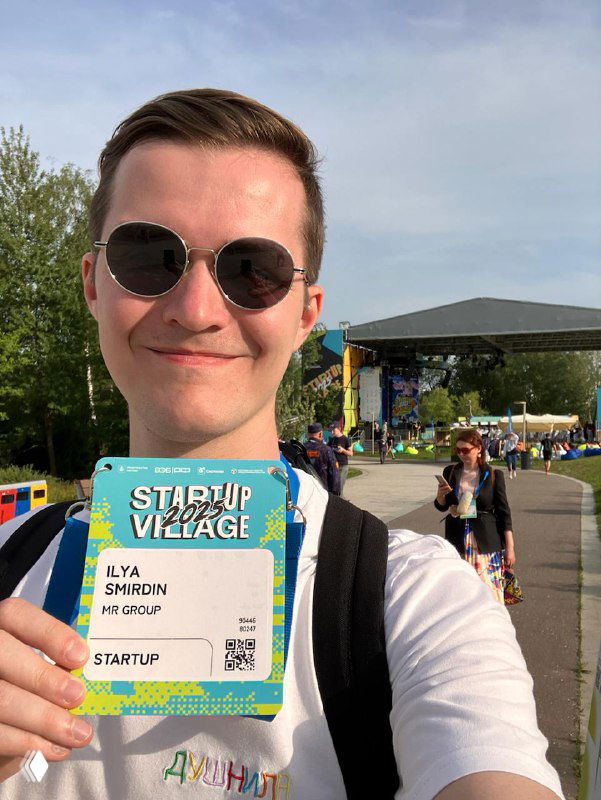 Автор держит бейдж Startup Village 2025 на открытой площадке в Сколково: сцена и люди на заднем плане в солнечный день, фестивальная атмосфера.