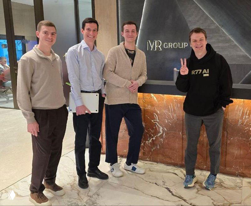 Четверо мужчин в деловой неформальной одежде в лобби на фоне логотипа R77 — встреча и нетворкинг после дискуссии.