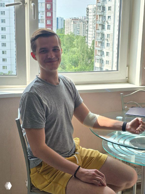 Автор сидит у окна с повязкой на руке после забора крови: виден городской пейзаж за окном и домашний интерьер на заднем плане, момент после анализа.