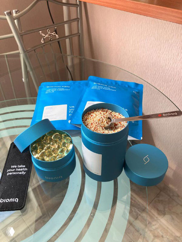 Распаковка набора Bioniq: синяя коробка, открытая баночка с гранулами, омега-3 в капсулах, ложечка и пакетики лежат на стеклянном столе.