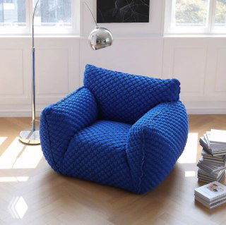 Синее объемное мягкое кресло с фактурной 3D‑тканью на светлом паркете у окна, рядом торшер и стопка журналов в интерьере