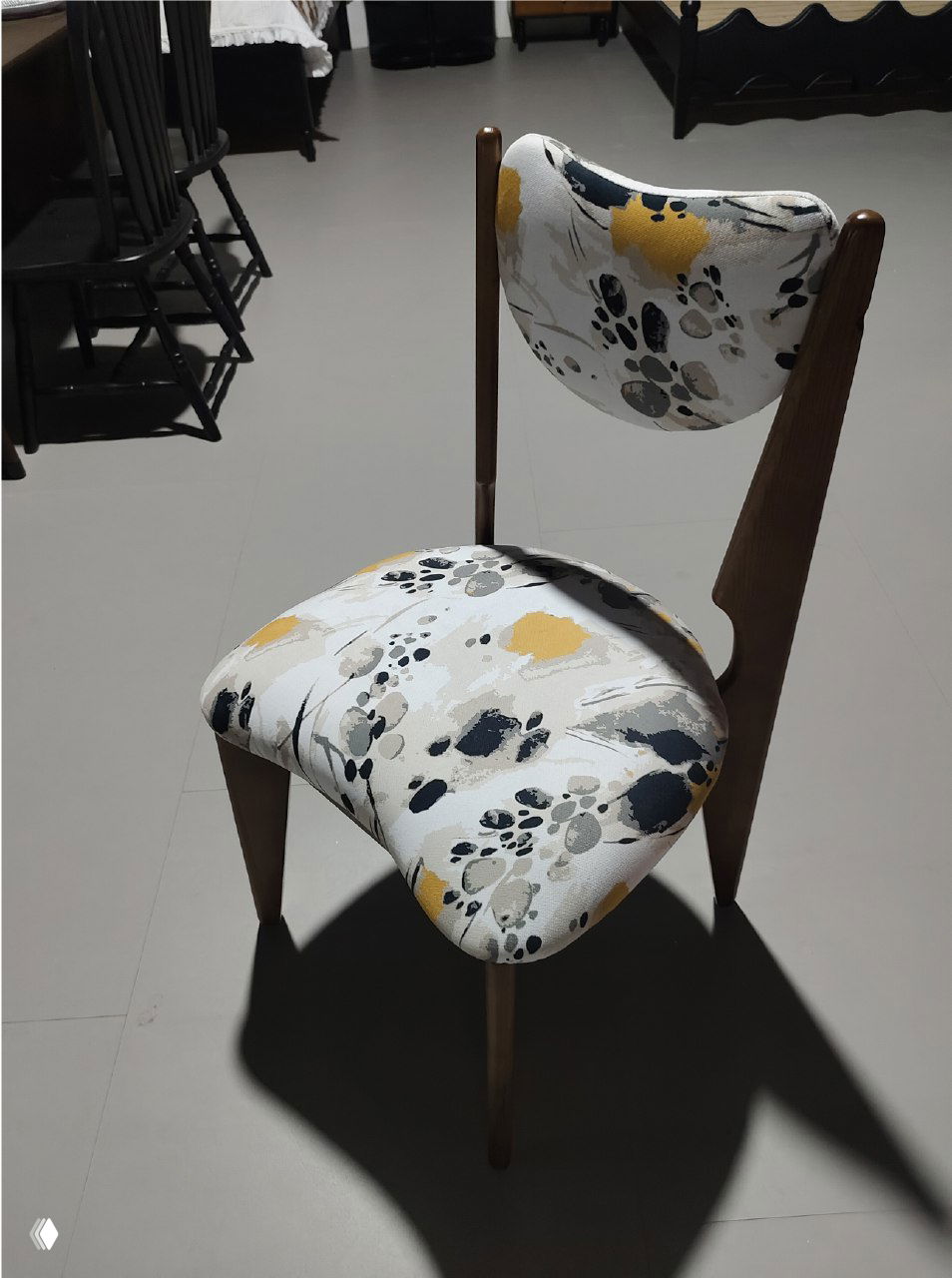 Фотография стула из массива ясеня с тканевой обивкой бело‑сине‑жёлтого абстрактного узора, показан в интерьере на светлом полу, вид под углом.