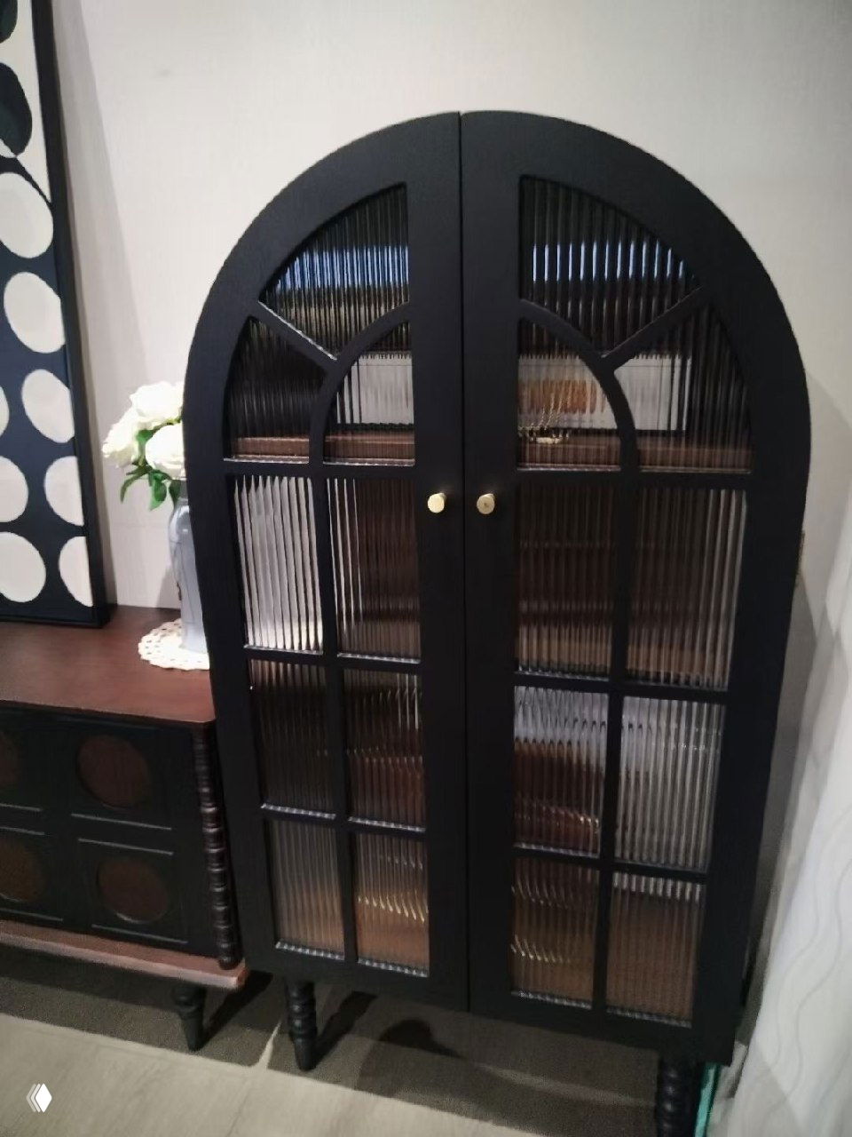 Чёрный арочный шкаф с рельефными стеклянными дверцами и декоративными ножками, установленный в жилом интерьере рядом с комодом и вазой с цветами.