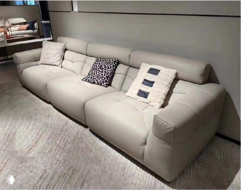 Широкий бежевый кожаный диван в шоуруме на сером ковре, три декоративные подушки и мягкая спинка, общий вид модели в интерьере.