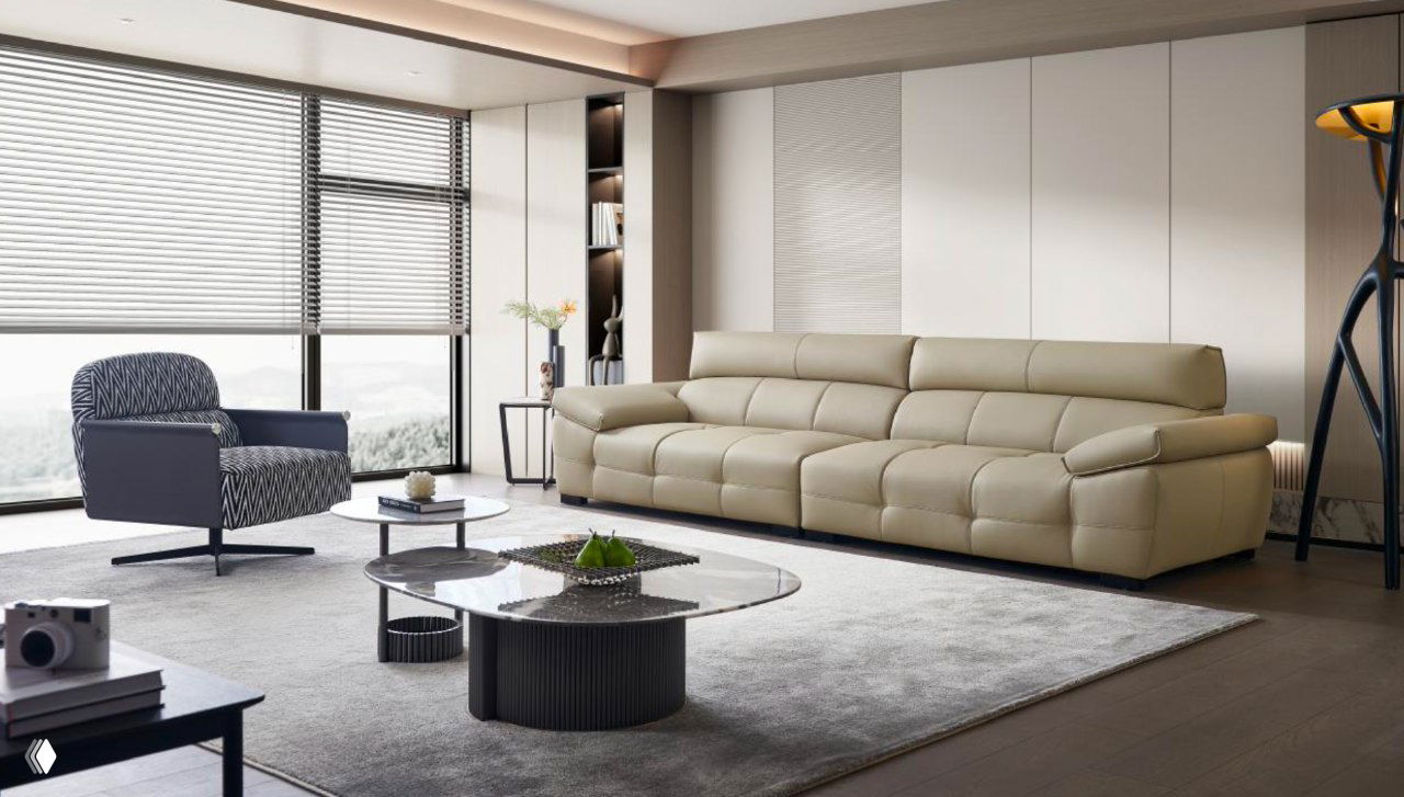 Светло‑бежевый кожаный трёхместный диван в просторной современной гостиной с журнальным столиком, креслом и панорамными окнами, показ профиля и отделки.