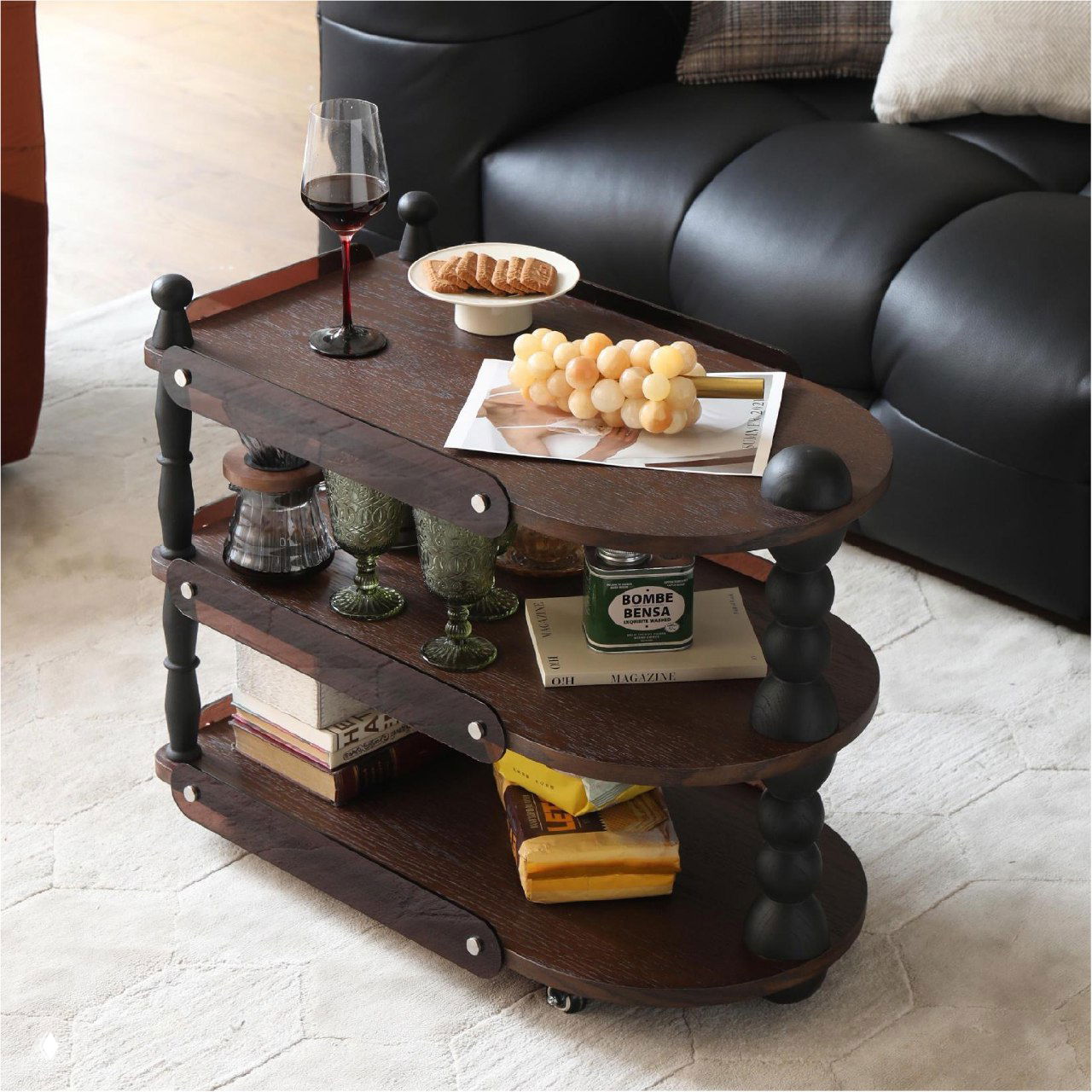 Трёхъярусный сервировочный столик из тёмного дуба на колёсиках в гостиной: бокал вина, печенье на тарелке, гроздь винограда, книги и декоративные стаканы у чёрного дивана