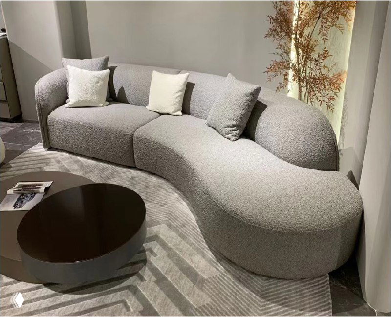 Серый изогнутый диван в выставочном интерьере с подушками и журнальным столиком, демонстрация формы и текстуры обивки.