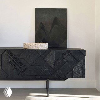 Крупный план фасада тумбы из ясеня с геометрическим резным узором и матовой чёрной отделкой на фоне светлой стены в комнате.