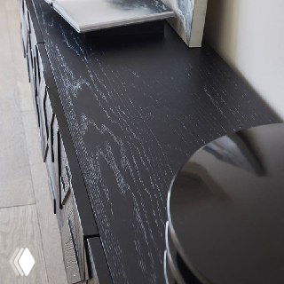 Верхняя поверхность тумбы из ясеня с видимыми волокнами древесины и матово-чёрной отделкой, предметы декора частично видны на столешнице.