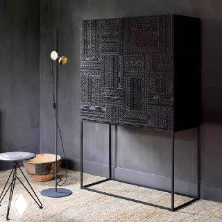 Угловой ракурс тёмного шкафчика из ясеня рядом с лампой и аксессуарами; вид подчёркивает боковой профиль, фактуру резного фасада и общую высоту изделия.