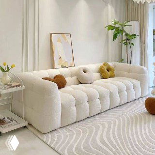 Прямой белый диван в жилой комнате с декоративными подушками и зелёным растением у окна, спокойная светлая обстановка и текстильные акценты