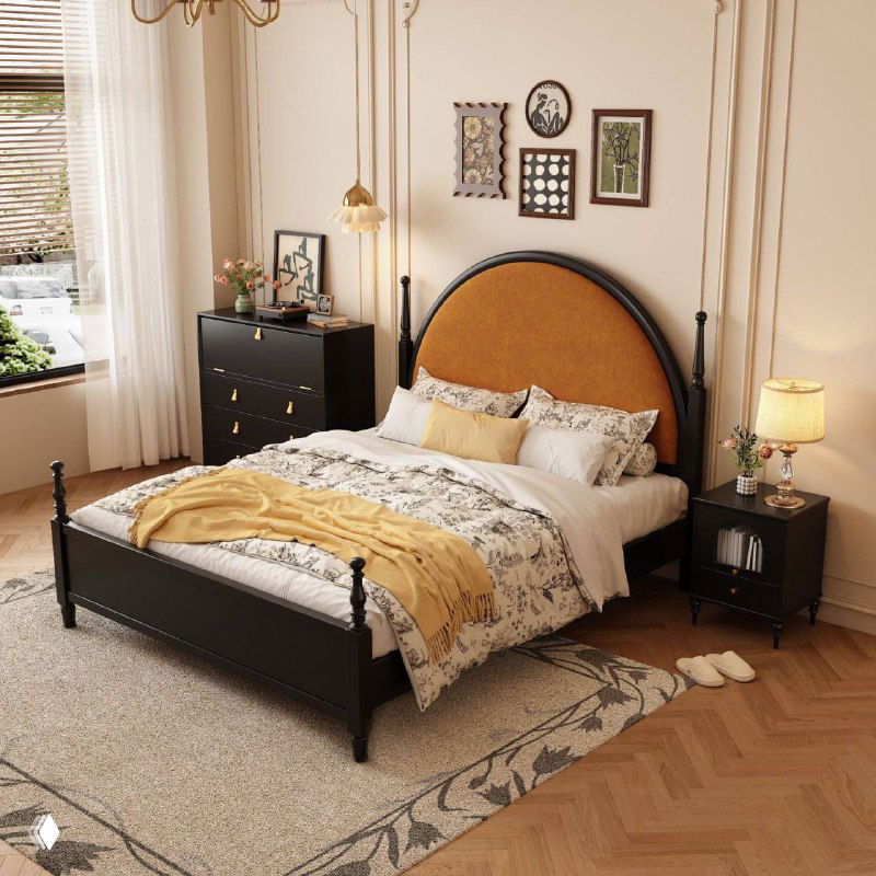 Вид кровати под углом: черная деревянная оправа, мягкое оранжевое изголовье, прикроватная тумба и лампа в просторной светлой комнате.