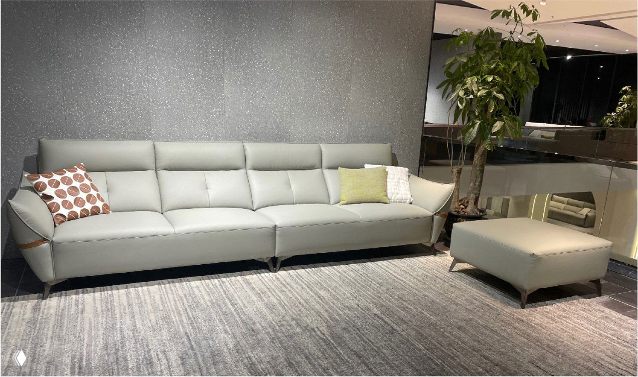 Широкий светло‑серый диван с пуфом в выставочном зале, современный минималистичный интерьер, декоративные подушки, ковер и большое растение рядом