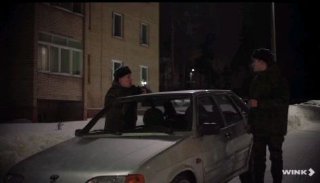 Ночное уличное фото: двое взрослых стоят у припаркованной легковушки возле жилого дома на заснеженной, заснеженной дороге; сцена выглядит напряжённой и атмосферной.