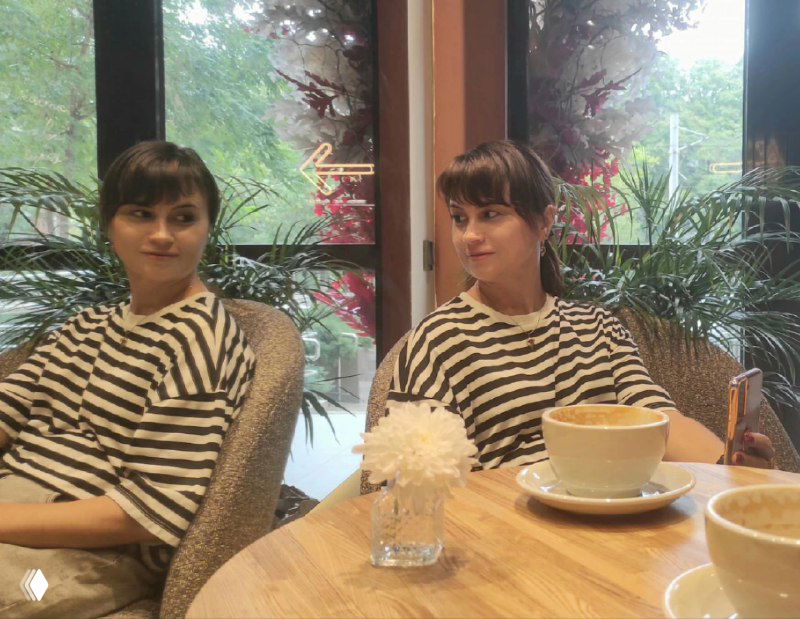 Женщина в полосатой кофте сидит за столом в кафе напротив зеркального отражения; на столе чашка кофе.