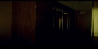 Кадр из фильма: тёмный коридор с деревянной отделкой, приглушённым боковым светом и глубокой тенью, создающий атмосферу напряжения сцены.