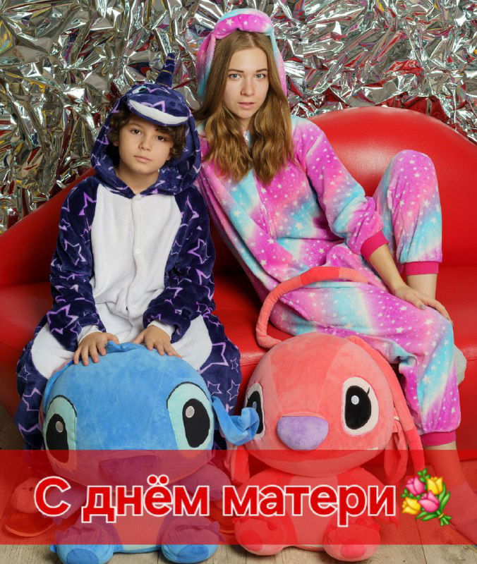 Две девочки/дети в ярких пижамах-кигуруми сидят на диване с мягкими игрушками; праздничная надпись «С днём матери» — тёплая семейная сцена.