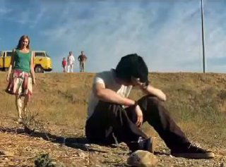 Кадр из фильма: ребёнок сидит на земле с опущенной головой, позади несколько взрослых и жёлтый фургон на холме — ощущение отчуждения и одиночества.