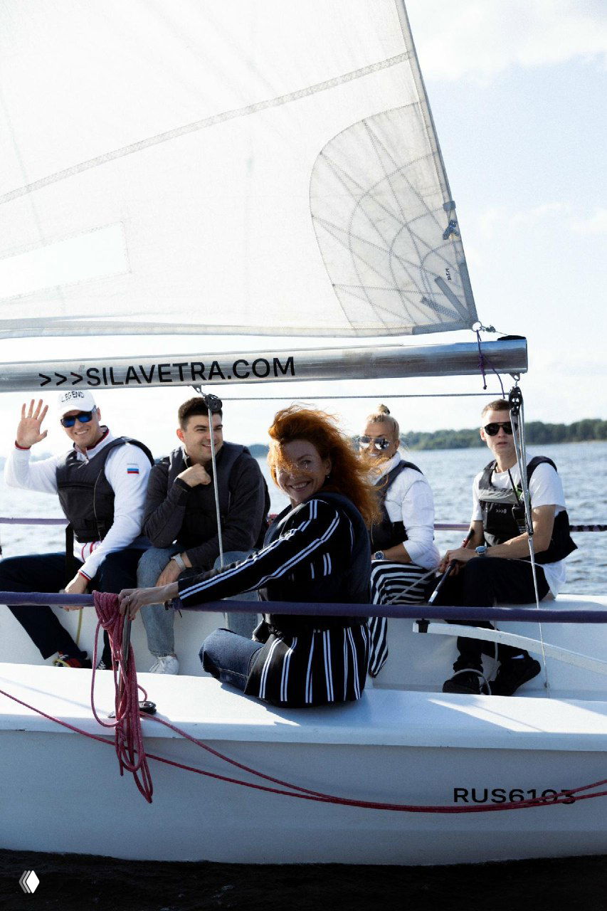 Гости на парусной прогулке: несколько человек в спасжилетах на борту яхты, видны парус и вода, участники смотрят в камеру и улыбаются.
