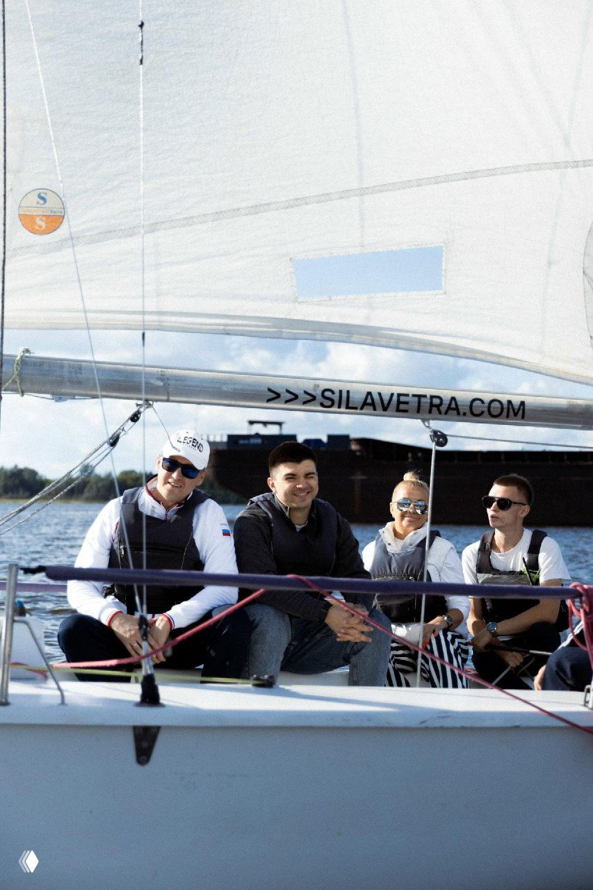 Группа гостей, расположившаяся на борту яхты под поднятым парусом; на переднем плане люди в солнцезащитных очках и спасжилетах, атмосфера прогулки.