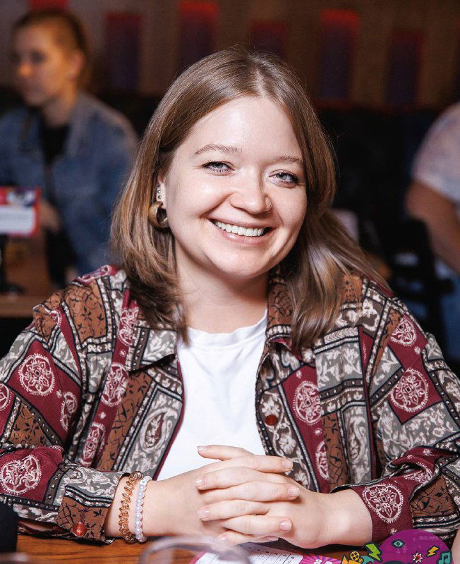 Улыбающаяся женщина в деловой обстановке сидит за столом; на заднем плане размытая аудитория, кадр передаёт тёплую рабочую атмосферу.