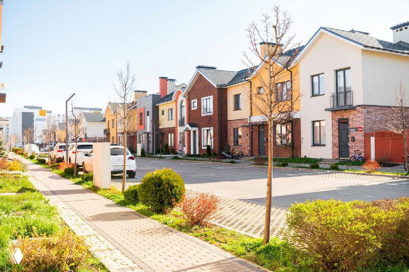 Фасады таунхаусов и коттеджей в аккуратном коттеджном поселке: солнечная улица, тротуар, газоны, молодые деревца и парковочные места на переднем плане.