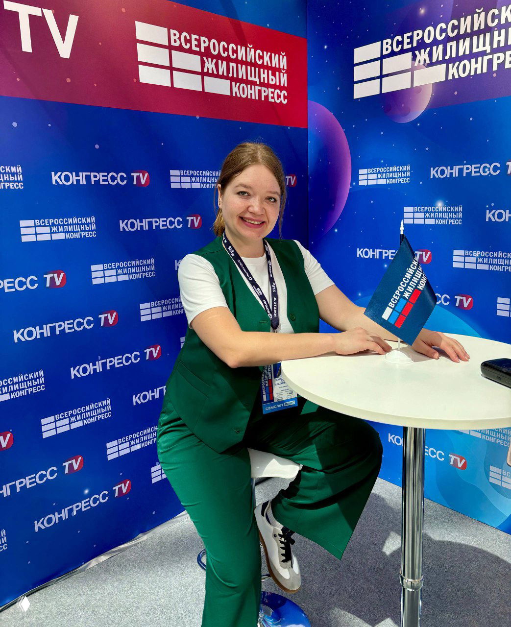 Женщина в зелёном костюме сидит за круглым столом и улыбается; на заднем плане баннер «Всероссийский жилищный конгресс», логотипы и флаг конференции, деловая обстановка