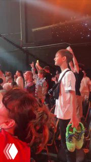 Мальчик в толпе на киберспортивном турнире, держит кроссовки в руках; вокруг яркие прожекторы, ликующие зрители и сцена финала.