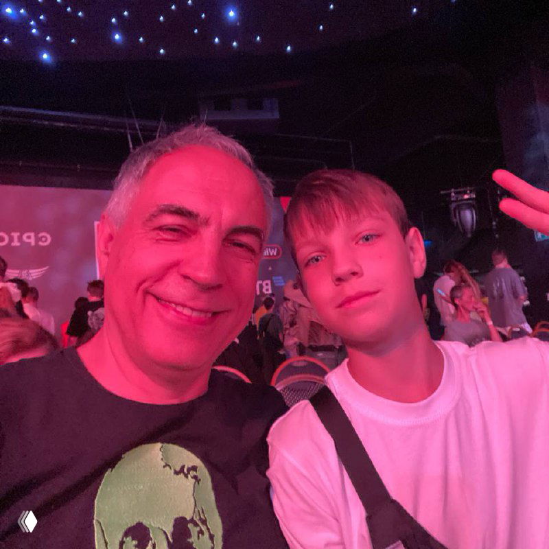 Отец и сын делают селфи в зале финала: их лица освещены розовой подсветкой, позади видна сцена, экран и суета фанатов.