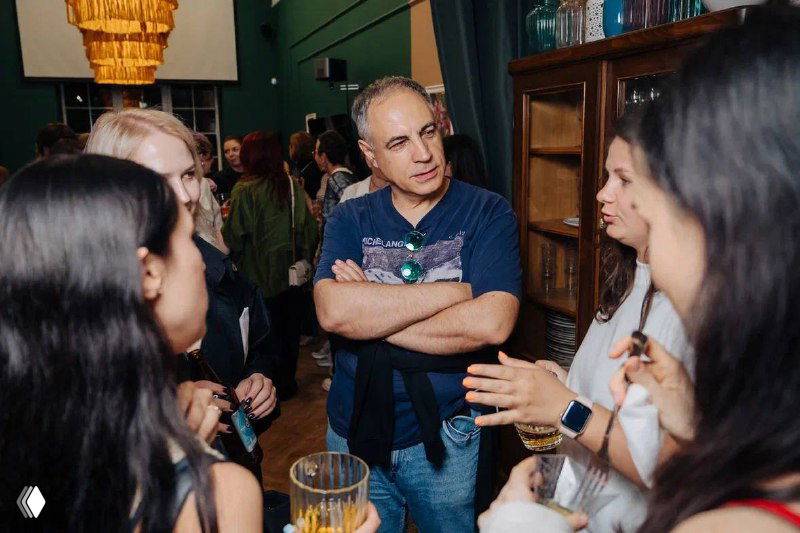 Группа людей в помещении общается: мужчина в центре с согнутыми руками говорит, вокруг несколько женщин слушают — беседа в непринуждённой атмосфере.