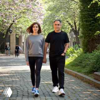 Мужчина и женщина средних лет идут по мощёной аллее в центре города, держатся за руки под зелёными кронами деревьев во время прогулки.