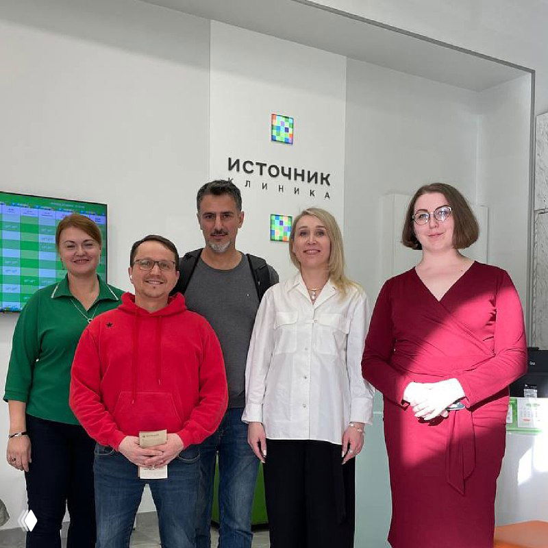 Группа сотрудников у стены с логотипом «Источник клиника» — фото команды и партнёров в приёмной, отражающее впечатление от пространства и корпоративной культуры учреждения.