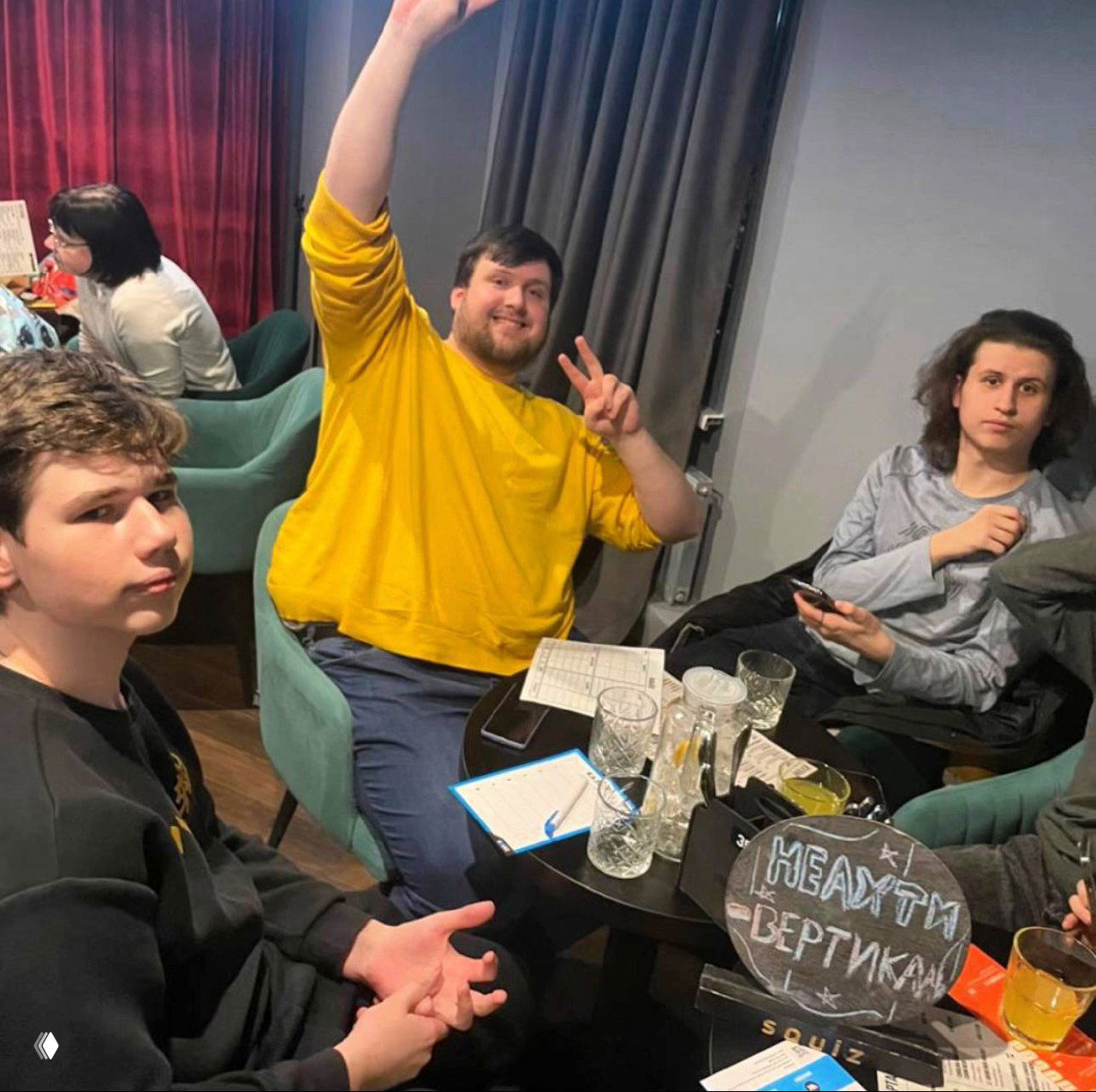 Группа подростков и преподаватель за столом с тетрадями, листами и напитками в непринуждённой обстановке обучения и общения.