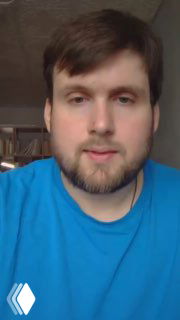 Портрет взрослого человека в голубой футболке в домашней обстановке, кадр из видеообращения автора, нейтральное выражение лица и мягкое освещение.