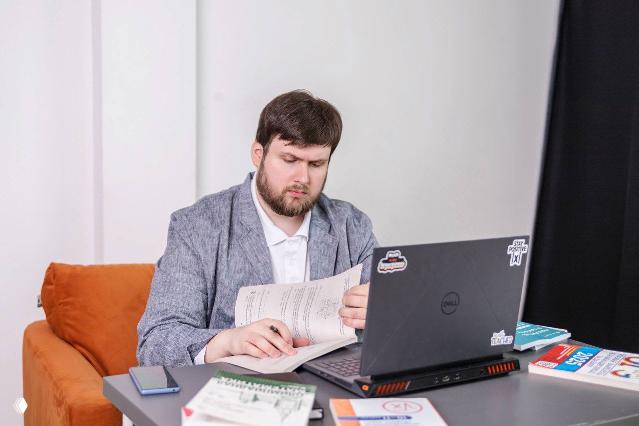 Мужчина за столом с ноутбуком и распечатками решает задания; на столе учебники и тетради — сцена подготовки к ОГЭ/ЕГЭ в рабочей обстановке.