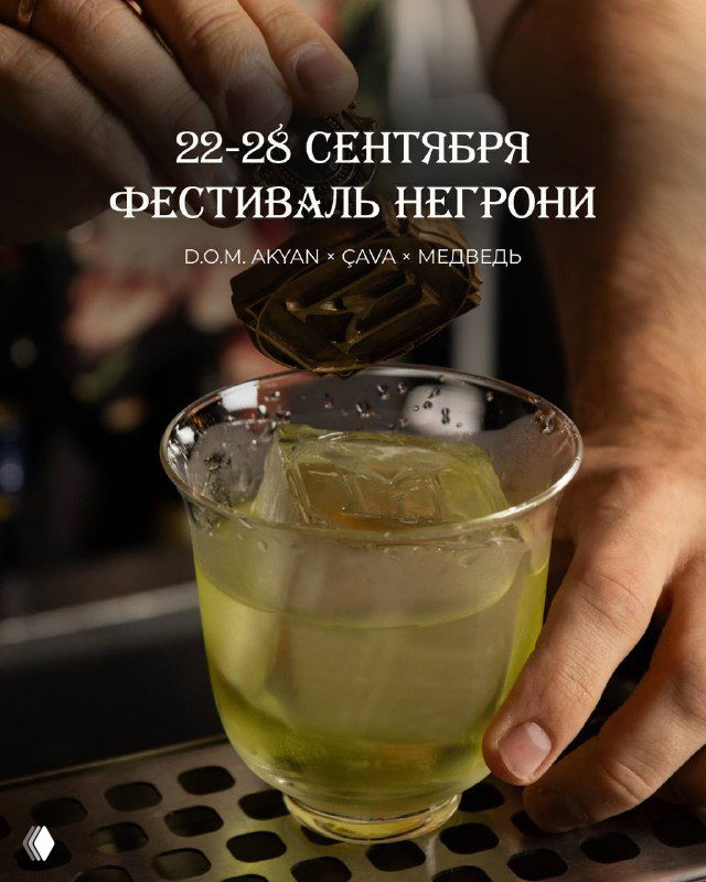 Рука держит бокал с бледно‑золотистым белым негрони Fleur d'or на барной стойке; видны крупные льдинки, решётка и размытый фон.