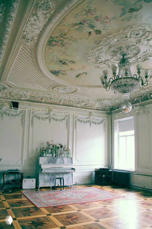 Интерьер парадной гостиной особняка: высокий потолок с фресками и лепниной, рояль у стены и большой ковер на деревянном полу.