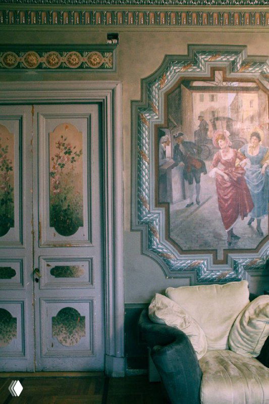 Декоративные панно и двери с росписью в историческом интерьере особняка, уютный диван у стены и изящные молдинги.