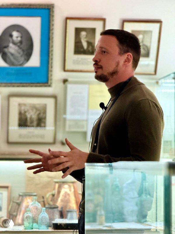 Лектор говорит перед витринами с музейными экспонатами на фоне портретов; кадр из авторской экскурсии о питьевой культуре.