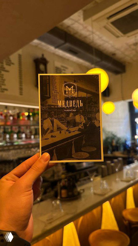 Рука держит карточку меню бара на фоне барной стойки с подсветкой — интерьер современного бара, где проходят встречи и экскурсии.