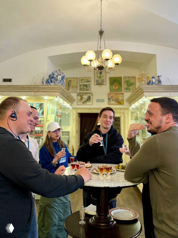 Группа участников стоит вокруг высокого столика с рюмками и беседует в зале музея или бара во время экскурсии‑лекции по истории алкоголя.