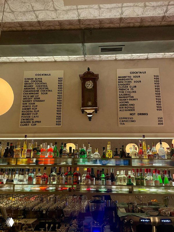 Барная стойка с полками бутылок и старинными часами на стене; на фоне видна большая доска-меню с перечнем коктейлей и цен в «Медведе».