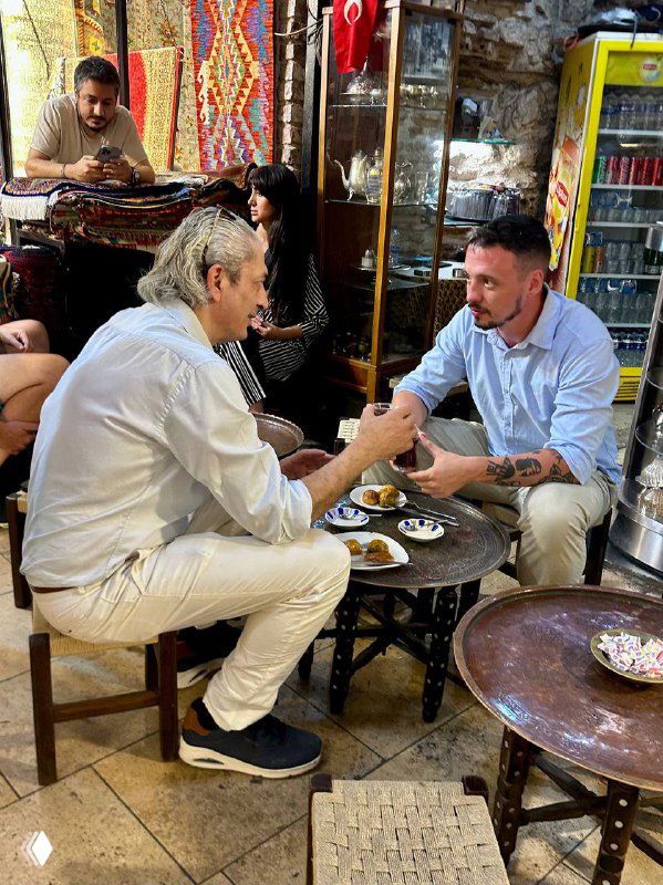 Двое мужчин сидят за низким столиком в проходной лавке/рыночной зоне, на столе стаканы турецкого чая Ince belli и небольшие тарелки с закусками, оживлённый фон рынка.