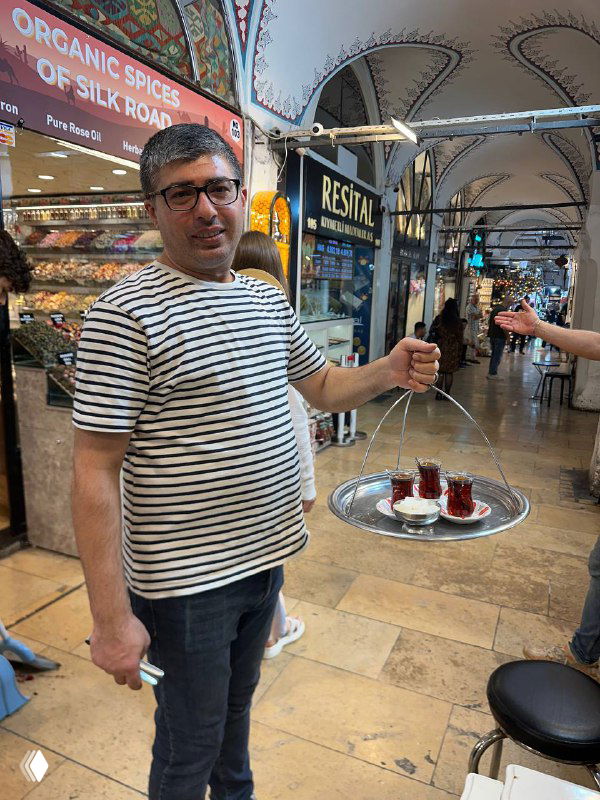 Продавец в полосатой футболке нес поднос с несколькими стаканами чая через крытую торговую галерею — типичная сцена подачи чая посетителям базара.