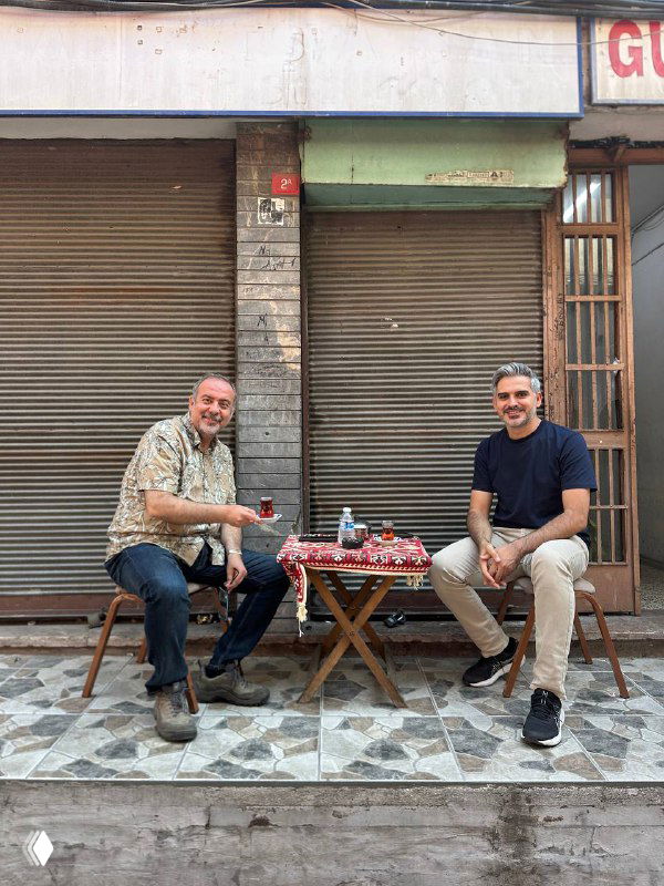 Двое мужчин сидят за маленьким столиком у закрытого магазина на тротуаре, перед ними стаканы крепкого турецкого чая и простая уличная обстановка.