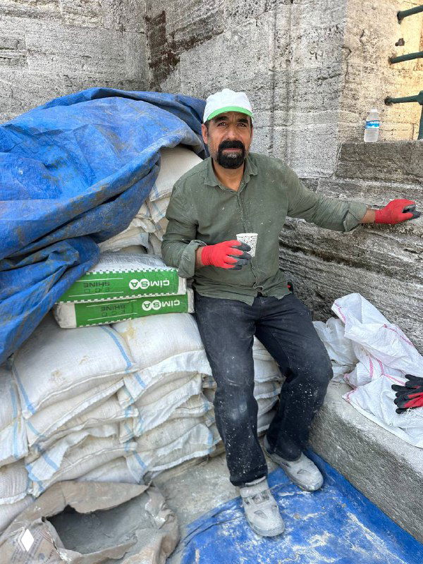 Рабочий в строительной или складской зоне делает паузу и держит стакан чая в руке — момент повседневного перерыва среди груды мешков и строительных материалов.