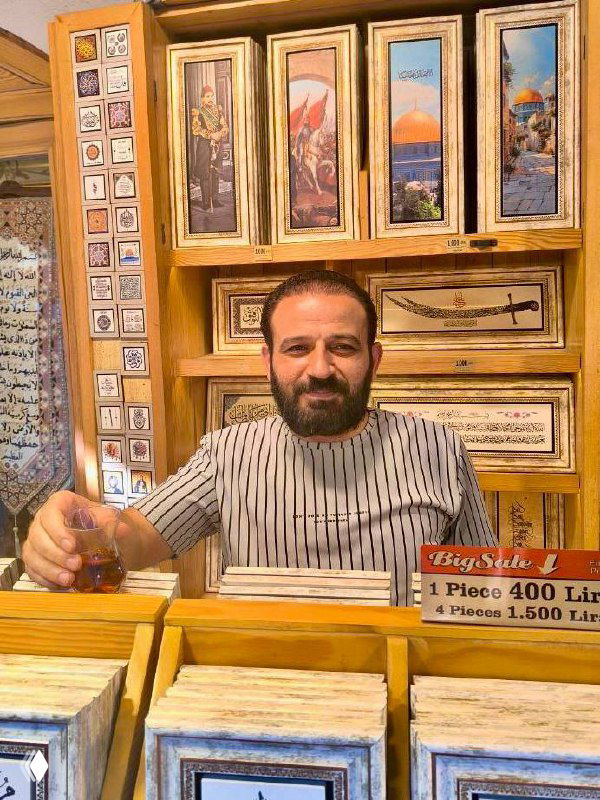 Продавец восточных сувениров или картин позирует за прилавком с чашкой чая — интерьер лавки с рамками и предметами искусства на заднем плане.