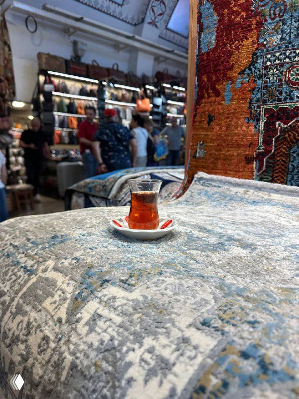 Одинокий стакан чая на столике, поставленном на покрытый ковром стол в магазине ковров; акцент на чашке и текстуре ковра вокруг.