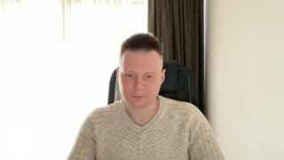 Портрет автора в светлом кабинете: мужчина в светлом пуловере сидит за столом на фоне занавески и кресла, нейтральное выражение лица