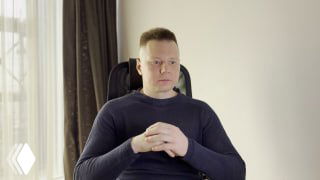 Портрет мужчины в офисе: сидит в кресле, руки сложены, серьезное нейтральное выражение, на фоне окно и шторы.